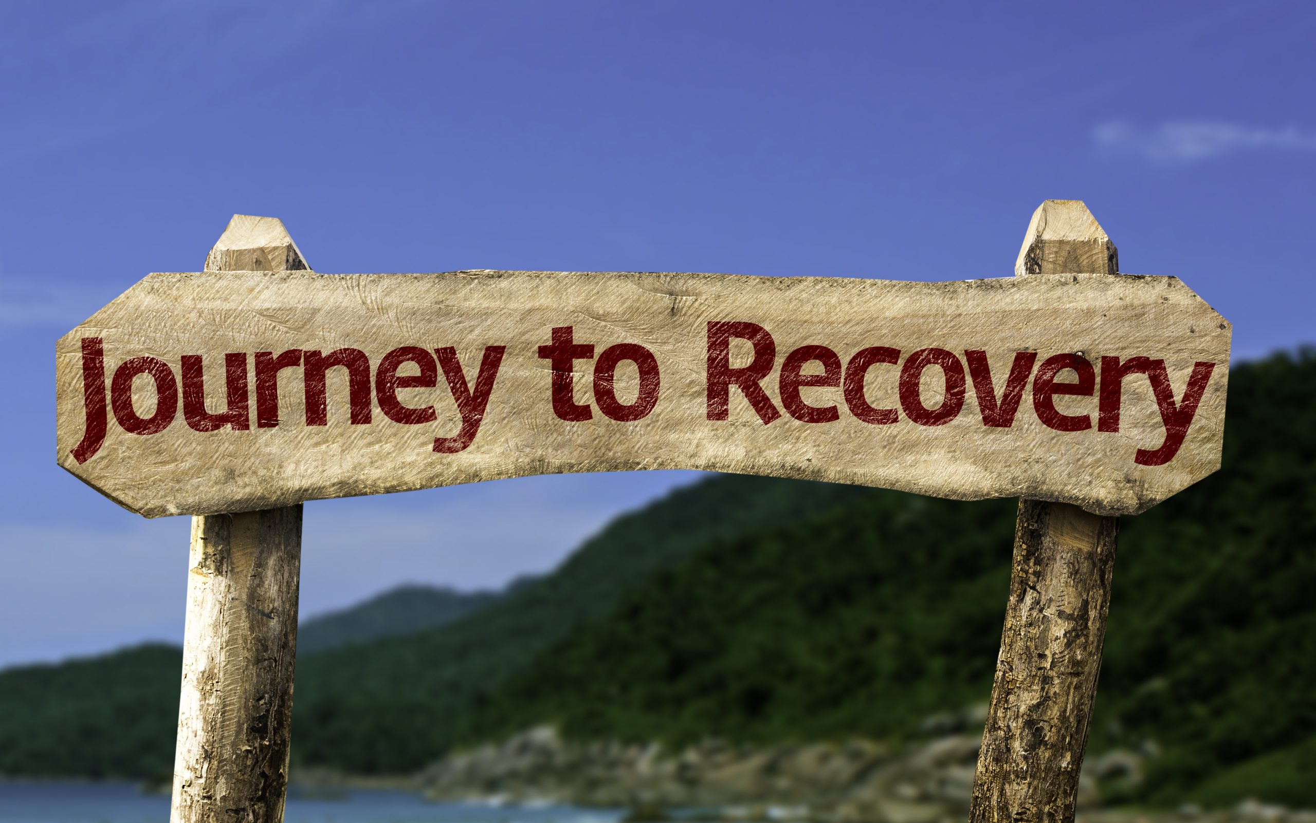 A sign reads: journey to recovery. The sign is outside with a hill behind it and blue sky.