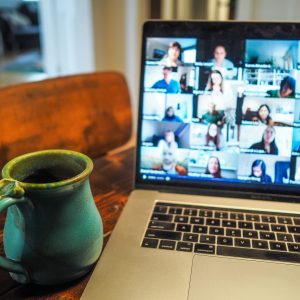 A coffee cup sits next to a laptop with many faces on a zoom chat.