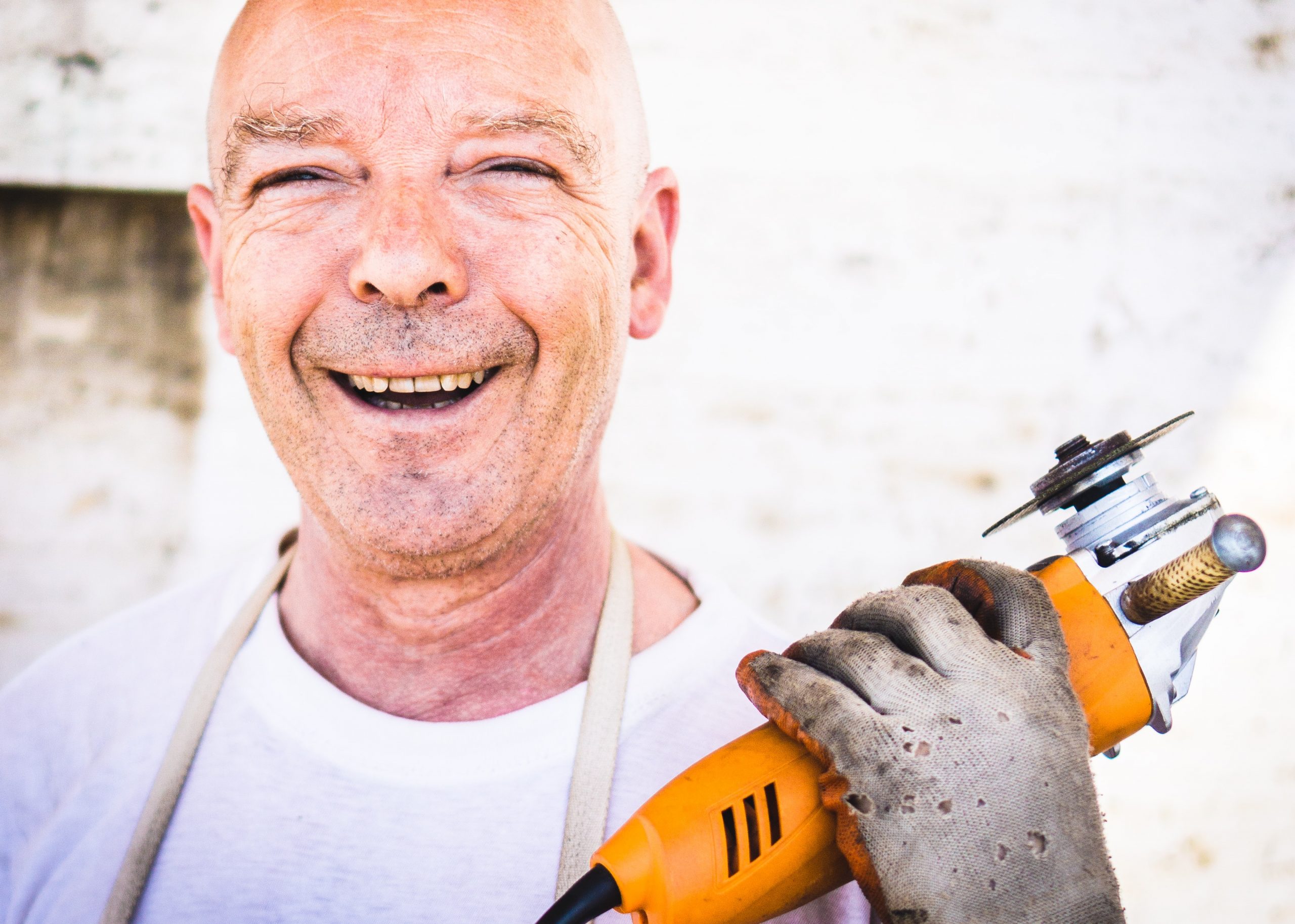 A man smiles, holding a drill and looks directly at the camera.