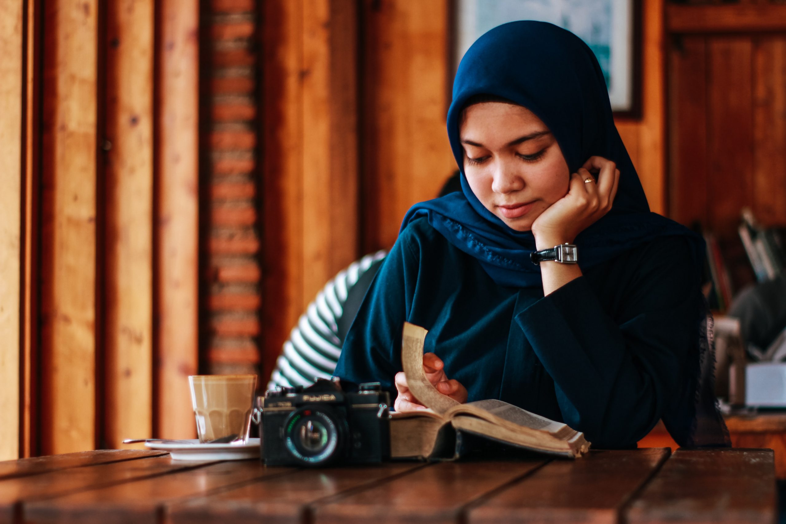 A woman sits at a table and contemplates. She wears a hijab.