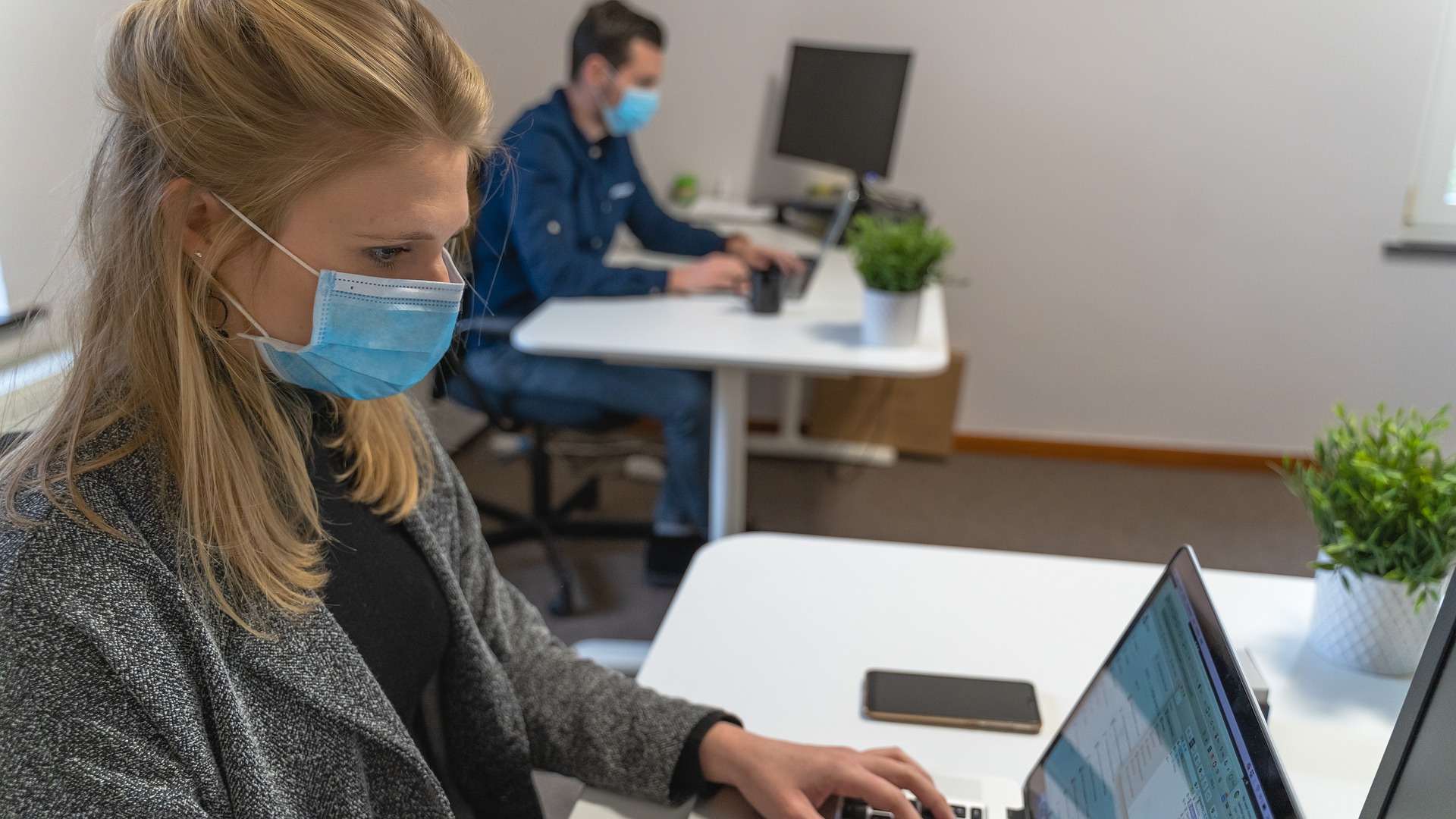 Two office workers work at desktop computers and wear masks.