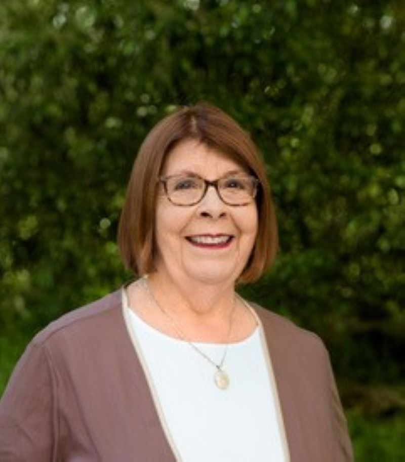 A lady stands in front of a green bush and smiles. She wears glasses.